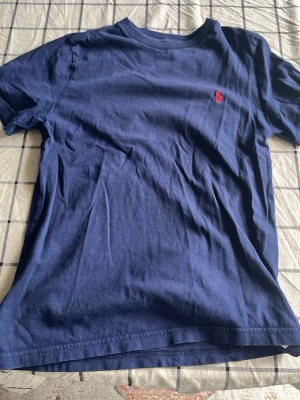 Mörkblå t-shirt från Polo Ralph Lauren - Mörkblå t-shirt från Polo Ralph Lauren med klassisk passform och korta ärmar. T-shirten har en liten röd broderad logga på bröstet och är tillverkad i mjuk bomull. Perfekt för en stilren och avslappnad look.