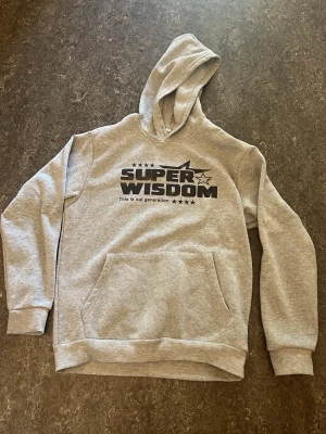 Grå hoodie med tryck SUPER WISDOM - Snygg grå hoodie med svart tryck på bröstet där det står 'SUPER WISDOM' och flera stjärnor. Hoodien har en stor känguruficka framtill, ribbade muddar och en huva med tryckt stjärna på baksidan. Perfekt för en avslappnad streetstyle-look. 