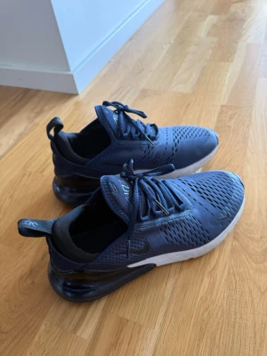 Blå Nike Air Max 270 sneakers - Säljer ett par blå Nike Air Max 270 sneakers med vit sula och svart detalj vid hälen. Skorna har snörning och en luftig ovandel i syntetmaterial. Ikonisk Air Max-bubbla i hälen för extra komfort och stil. Använda bara 1 gång. Helt nya.