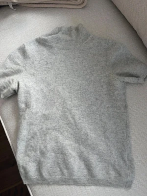 Ljusgrå stickad t-shirt Soft Goat - Ljusgrå stickad t-shirt från Soft Goat i 100% kashmir. Tröjan har korta ärmar, ribbad hög hals och ribbade muddar vid ärmslut och nederkant. Passar bättre på en XS än S