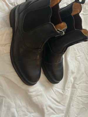 Svarta Chelsea boots i läder - Säljer ett par klassiska svarta Chelsea boots i äkta läder med elastiska sidopaneler och låg klack. Insidan är fodrad i ljusbrunt material och bootsen har en stilren, tidlös design som passar till många outfits.