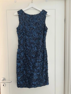 Blå paljettklänning med blommönster - Snygg blå kortklänning med blommig spets och rund halsringning. Klänningen är ärmlös och har en djupare rygg. Perfekt för dig som vill sticka ut med en elegant look. Materialet är troligtvis polyester eller liknande syntet. Klänningen är omsydd och använd ett fåtal gånger passar en storlek 36.