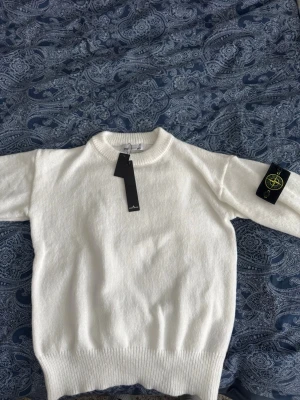 Vit stickad tröja från Stone Island - Vit stickad tröja från Stone Island med klassisk rund halsringning och ribbade muddar. Tröjan har den ikoniska Stone Island-loggan på vänster ärm och är tillverkad i mjukt material som ger en clean och stilren look. Priset kan diskuteras och det är storlek L men passar M och S. 