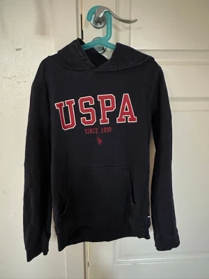 Svart hoodie från USPA med tryck - Svart hoodie från USPA med stor röd och vit logga på bröstet och texten 'Since 1890'. Klassisk känguruficka framtill och huva. Perfekt för en avslappnad och sportig stil.