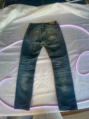 Blå jeans från Nudie Jeans - Säljer ett par klassiska blå jeans från Nudie Jeans med snygga slitningar och raka ben. Jeansen har fem fickor och detaljerade sömmar på bakfickorna. Perfekta för dig som gillar en avslappnad och tidlös stil. straight/bootcut