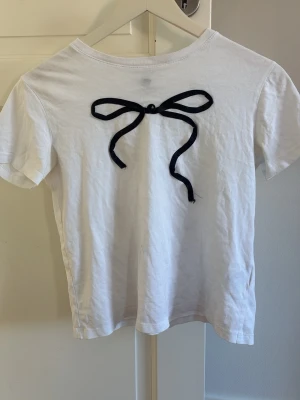 Vit t-shirt med svart rosett - Vit t-shirt i bomull med en svart rosettdetalj framtill. Klassisk rund halsringning och korta ärmar. Perfekt för en stilren och söt look.