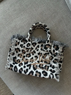 Leopardmönstrad shoppingväska - Säljer en leopardmönstrad shoppingväska med fransade kanter och texten 'Summer Vibes' broderad på framsidan. Väskan har två handtag, dragkedja och innerfack. Materialet är tyg och färgerna går i beige, svart och brunt. Perfekt för dig som gillar statement-accessoarer.