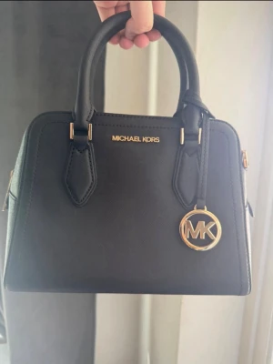 Michael Kors väska - Stilren svart handväska från Michael Kors i strukturerat läder med guldfärgade metalldetaljer och logotyp. Väskan har dubbla handtag, dragkedja och en rund MK-berlock. knappt använd!