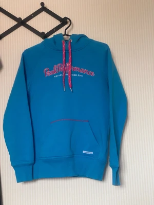 Blå hoodie från Peak Performance - Snygg blå hoodie från Peak Performance med rosa snören och broderad logga på bröstet. Hoodien har magficka, ribbade muddar och huva. Perfekt för en avslappnad och sportig stil.