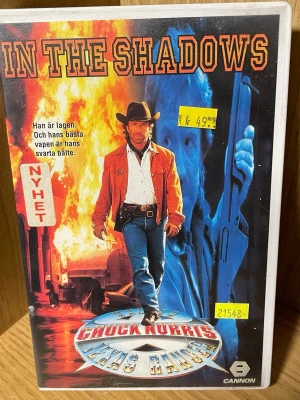 Walker Texas Ranger VHS: In The Shadows - VHS-film med titeln 'In The Shadows' från serien Walker Texas Ranger. Omslaget har en actionfylld design med eld, blåa och orange färger samt tydlig Texas Ranger-tema. Text på svenska och klassisk VHS-förpackning i plast.