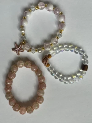 3-pack pärlarmband i pastellfärger - Säljer tre elastiska armband med pärlor i pastellrosa, vitt och transparent. Ett armband har en sjöstjärneberlock och gulddetaljer, ett har en rosformad dekoration och träpärlor, och det tredje har glittriga rosa pärlor. Perfekt för att ge din stil en söt touch.