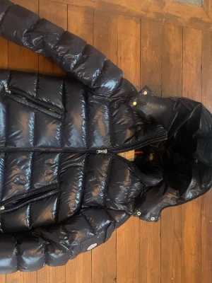 Mörkblå  Moncler pufferjacka med huva - Säljer en Mörkblå pufferjacka från Moncler med glansig finish och stor huva. Jackan har två snedställda fickor med dragkedja, quiltad design och Moncler-logga på ärmen. Perfekt för kalla dagar och har en riktigt schysst streetstil. Skriv vid funderingar eller prisförslag! Finns ett litet hål i vänstra armhålan som knappt syns,går enkelt att sy ihop!