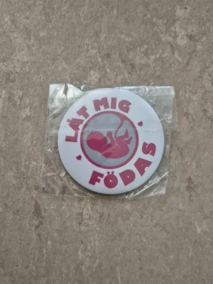 Vit knapp med text 'Låt mig föd - Rund vit klassisk knapp med nål på baksidan. Knapp med rosa texten 'Låt mig födas' och en illustration av ett foster i mitten. Knappen har säkerhetsnål på baksidan och är tillverkad i metall och plast. Perfekt för att fästa på jacka eller väska.