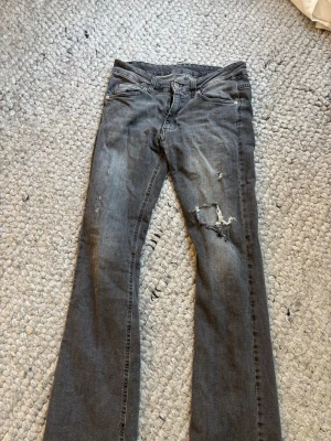 Grå slitna bootcut jeans - Säljer ett par grå bootcut jeans med slitningar och hål på ena benet. Jeansen har en låg midja. Jag vet att jag köpte de från kidsbrandstore men kommer inte ihåg vilket märke