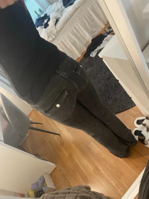 Svarta bootcut jeans med fickdetaljer - Säljer ett par svarta bootcut jeans med snygga fickor bak som har lock och silverfärgade knappar. Jeansen har klassisk femficksdesign och är i stretchigt material för skön passform. Perfekta till en avslappnad stil.