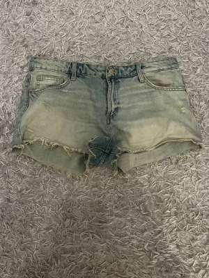 Ljusblå jeansshorts med fransig kant - Säljer ett par ljusblå low waist jeansshorts med fransig och rå kant nertill. Shortsen har klassisk femficksdesign, knapp och dragkedja framtill samt bälteshällor. Perfekta för varma dagar och en avslappnad stil.