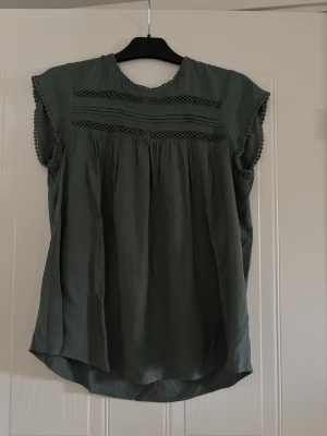Grön blus med spetsdetaljer - Snygg grön blus med rund halsringning och dekorativa spetsdetaljer framtill. Blusen har korta ärmar med små bollar längs kanterna och ett luftigt, löst fall. Perfekt för en avslappnad men stilren look.