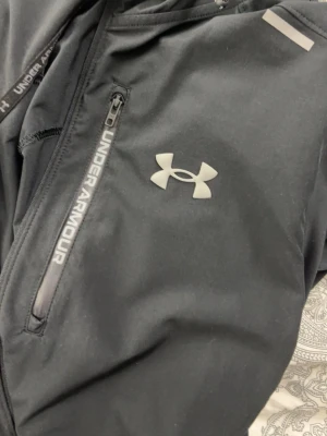 Svart vindjacka från Under Armour - Svart vindjacka från Under Armour med huva och dragkedja framtill. Jackan har logga på bröstet och detaljer med Under Armour-tryck längs dragkedjan och nederkanten. Perfekt för aktiva dagar och sportiga outfits.