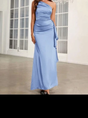 Ljusblå one shoulder långklänning - Elegant ljusblå långklänning med one shoulder-design och draperad detalj i midjan. Klänningen har en slät, glansig yta och en figurnära passform upptill som faller mjukt nedåt. Perfekt för speciella tillfällen.