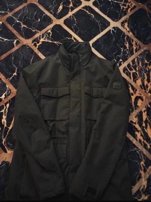 J.Lindeberg Jacka (field) - Stilren svart vindjacka med hög krage och flera praktiska fickor framtill. Jackan har en diskret logotyp på ärmen och stängs med dragkedja. Perfekt för dig som gillar en clean och funktionell look.