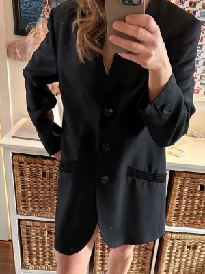 Fin Svart oversized kavaj från Gerry Weber Stl 38  - Stilren svart oversized kavaj från Gerry Weber med tre knappar framtill och två fickor med dekorativa detaljer. Rak passform och utan krage för en modern look. Perfekt att styla till olika outfits för en trendig vibe.