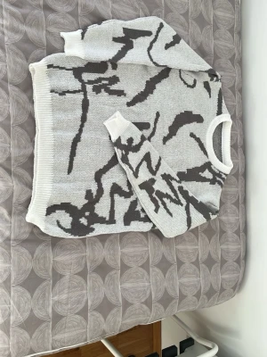 Vit stickad tröja med svart mönster - Snygg vit stickad tröja med abstrakt svart mönster. Tröjan har rund halsringning och ribbstickade muddar vid ärmslut och nederkant. Perfekt för dig som gillar unik design och vill sticka ut lite extra.