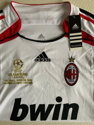 AC Milan Tröja  - Vit AC Milan matchtröja från Adidas med svarta och röda detaljer, broderat Champions League-märke och klubbemblem. Stort Bwin-tryck på bröstet och guldtext om finalen i Aten 2007. Tillverkad i lätt och ventilerande material, perfekt för fotbollsträning eller samling.