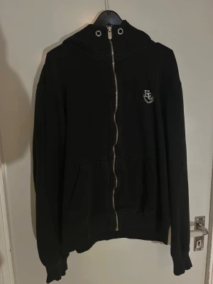 Svart zip hoodie med logga ETIREL - Svart hoodie med dragkedja från ETIREL. Broderad vit logga på bröstet och stora fickor framtill. Klassisk huva utan snöre och ribbade muddar vid ärmslut och midja. Perfekt för en avslappnad streetwear-look.