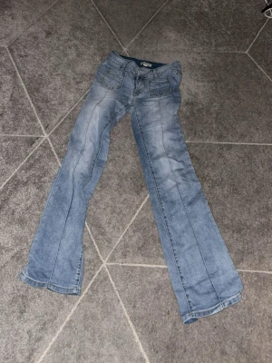 Ljusblå bootcut jeans med fickor - Säljer ett par ljusblå bootcut jeans med snygga slitningar och framfickor. Jeansen har även två bakfickor med lock och knappdetalj. Klassisk femficksmodell i mjukt denimtyg, perfekta för en avslappnad stil.