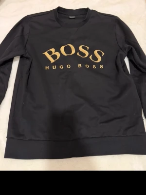 Svart sweatshirt från Hugo Boss - Snygg svart sweatshirt från Hugo Boss med stort guldfärgat BOSS-tryck på bröstet. Klassisk rund halsringning och ribbade muddar vid ärmar och nederkant. Perfekt för en stilren och avslappnad look. Storlek L passar På M 