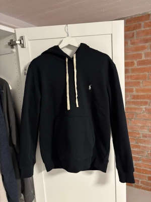 Svart hoodie från Polo Ralph Lauren - Svart hoodie från Polo Ralph Lauren med vit broderad logga på bröstet och vita dragsnören. Använd fåtal gånger och i fint skick. Storlek L men hade passat en M bättre. Priset är ktne hugget i sten så hör av er vid minsta fundering!