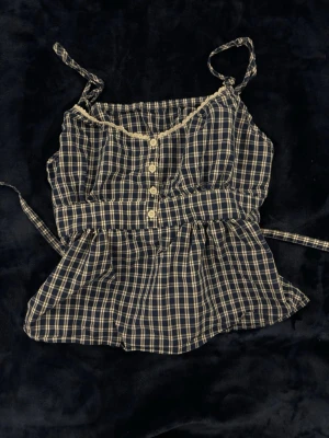 Rutig blå linnetopp med knytband - Supersöt rutig linnetopp i blått och beige med tunna axelband som knyts. Toppen har knappar framtill, volangkant nedtill och smock i ryggen för skön passform. Perfekt för sommarens alla tillfällen och ger en chill vintage vibe. Köpt i Shanghai på W managment, i storlek S men storlekar i Asien är mindre så passar bättre XS/XXS