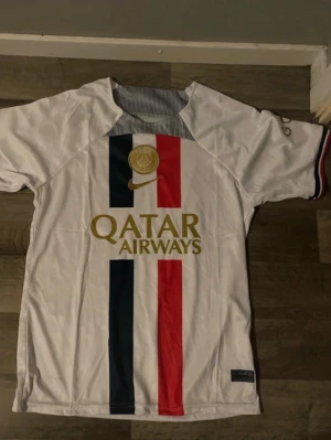 PSG matchtröja Nike vit  - Säljer en vit PSG matchtröja från Nike med Qatar Airways-tryck i guld. Tröjan har vertikala ränder i mörkblått och rött framtill samt PSG-logga och Nike swoosh i guld. Perfekt för fotbollsfans som vill sticka ut på planen eller läktaren.