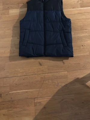 Väst - Snygg mörkblå dunväst med quiltat mönster och två fickor framtill. Västen har en hög krage och stängs med dragkedja. Perfekt för lager-på-lager och håller dig varm under kyliga dagar.