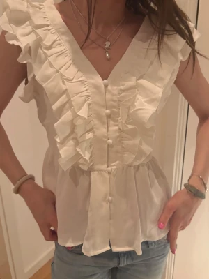 Vit volangblus med knappar - Söt vit blus med volangdetaljer längs axlar och bröst, samt dekorativa knappar framtill. Blusen har v-ringning och är ärmlös, vilket ger en luftig och trendig look. Perfekt att matcha med jeans eller kjol för en fräsch stil.