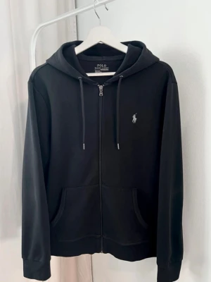 Svart hoodie från Polo Ralph Lauren - Säljer en svart hoodie från Polo Ralph Lauren med dragkedja framtill och klassisk vit logga på bröstet. Hoodien har huva med dragsko och två fickor fram. Tillverkad i mjuk bomullsblandning, perfekt för en avslappnad stil.