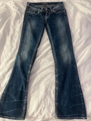 Blå bootcut jeans från Silver Jeans - Säljer ett par snygga blå bootcut jeans från Silver Jeans med slitningar och kontrastsömmar. Jeansen har klassiska fem fickor och broderade bakfickor med S-detalj. Tillverkade i stretchigt denim för skön passform. Jeansen har även små små blush fläckar. köpte dessa på plick men bara använt 1 gång. 
