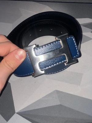 Blått skärp med silverfärgat H-spänne - Snyggt blått skärp från Hermès med ett stort H-format spänne i silverfärg. Skärpet har vita sömmar längs kanterna och är gjort i skinn med en stilren och modern look. Perfekt accessoar för att lyfta din outfit.