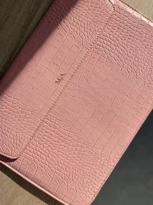 Rosa datorfodral i krokopräglat skinn - Snygg rosa kuvertväska med krokopräglat mönster och stilren design. Väskan har en enkel klaffstängning och initialerna M.A. på framsidan. Perfekt accessoar för att lyfta din outfit med en trendig touch.