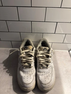 Vita Nike Air Force 1 sneakers - Klassiska vita Nike Air Force 1 sneakers i lågt utförande. Skorna har perforerad tå, tjock sula och Nike-logga på sidan samt tungan. Tillverkade i läder med textildetaljer och vita skosnören. Perfekta för en streetwear-look.