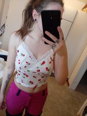 Vit croppad linnetopp med körsbär - Söt vit croppad linnetopp med körsbärsmönster och spetsdetaljer vid halsen. Tunn och lätt med smala axelband, perfekt för varma dagar. Toppen har omlott-design framtill och sitter tight mot kroppen.