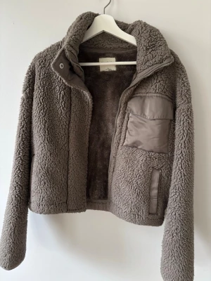 Brun teddyjacka Abercrombie & Fitch - Mysig brun teddyjacka från Abercrombie & Fitch Soft A&F Collection. Jackan har hög krage, dragkedja, en bröstficka och detaljer i blankt tyg. Perfekt för kalla dagar och ger en trendig look med sin fluffiga textur.