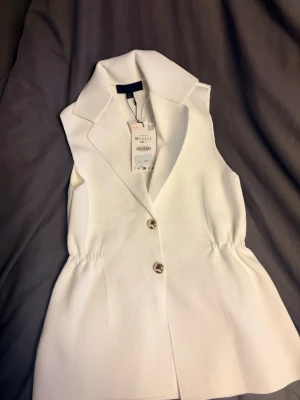 Vit väst med knappar från Zara - Stilren vit väst från Zara med två knappar framtill och markerad krage. Västen har en lätt insvängd midja och är tillverkad i ett strukturerat tyg som ger en snygg look. Perfekt att styla över en skjorta eller t-shirt för en trendig outfit.