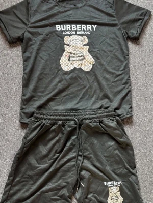Svart Burberry set med björntryck - Snyggt svart set från Burberry med t-shirt och shorts. Båda plaggen har ett mönstrat björntryck och texten 'BURBERRY LONDON ENGLAND' framtill. Setet är gjort i mjukt material och har resår i midjan på shortsen för extra komfort.