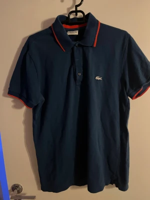 Marinblå pikétröja från Lacoste - Säljer en marinblå pikétröja från Lacoste med klassisk krage och orangea detaljer på krage och ärmslut. Tröjan har korta ärmar, knappar framtill och den ikoniska krokodilloggan på bröstet. Regular fit och tillverkad i bomull. Storlek 4 vilket är medium.
