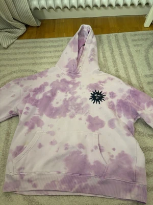 Lila tie-dye hoodie från SWEET SKTBS - Säljer en oversized hoodie från SWEET SKTBS i lila och vit tie-dye. Stor huva, magficka och svart soltryck på bröstet. Stor SWEET SKTBS-text på ryggen. Perfekt för en chill och streetwear-inspirerad look.
