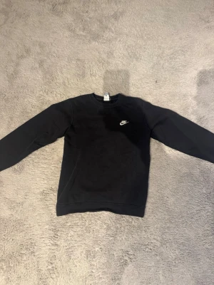 Svart crewneck från Nike - Svart sweatshirt från Nike med klassisk rund halsringning och vit broderad logga på bröstet. Tröjan har långa ärmar och ribbade muddar vid ärmslut och nederkant. Perfekt för en chill och sportig stil.