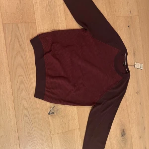 Vinröd tröja från BOSS - Vinröd tröja från BOSS med rund halsringning och ribbade muddar. Tröjan har ett diskret mönster på framsidan och är gjord i mjukt material, 74 virgin wool och 26 cotton. Helt ny och oanvänd, lappen är kvar och påsen den skickades i. Storleken var dock fel, därav ingen bild på. 