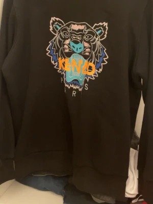 Svart Kenzo sweatshirt med tiger - Svart sweatshirt från Kenzo med ikoniskt broderat tigerhuvud i blått, rosa och turkost samt orange Kenzo-text på bröstet. Storlek L men skulle säga att den passar M