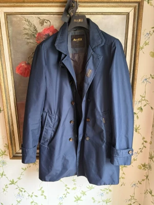 MooRER rock - Snygg mörkblå rock från MooRER Verona med klassisk krage och dubbelknäppning. Rocken har två sidofickor och detaljer som diskret logotyp på bröstet. Materialet är slätt och vattenavvisande, perfekt för stilrena dagar. Väldigt bra skick. Nypris är 10000kr+. Passar som M. 